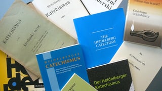 Mehrere Ausgaben der Heidelberger Katechismus, deutsche und fremdsprachige, liegen auf einem Tisch angeordnet.