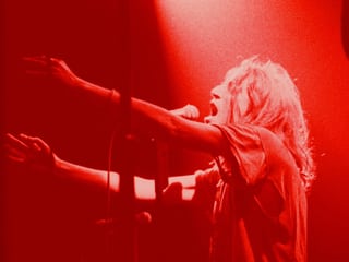 In rotem Licht steht die Sängerin Patti Smith auf der Bühne und hält die Arme nach vorne gestreckt.