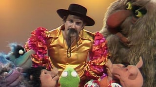John Cleese umgeben von den Muppets. Er trägt ein Mexikaner-Kostüm.