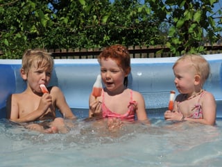 Aufblasbares Schwimmbecken für Kinder. Es sitzen 3 Kinder mit Glace im Wasser. Es scheint heiss zu sein und die Sonne scheint. 