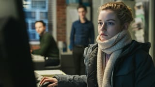 Eine Frau sitzt an einem Bürotisch, im Hintergrund schauen ihr zwei Männer zu Eine Frau sitzt an einem Bürotisch, im Hintergrund schauen ihr zwei Männer zu