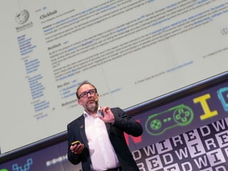 Ein Mann mit Brille und schwarzem Sakko steht vor einer Leinwand und erklärt etwas. Ein Mann mit Brille und schwarzem Sakko steht vor einer Leinwand und erklärt etwas.