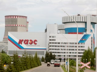 Zu sehen ist ein russisches Atomkraftwerk mit zwei  Kühltürmen. Davor stehen drei Personenwagen und das Werk ist mit kyrillischer Schrift bemalt.