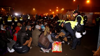 Flüchtlinge sitzen am Boden, Polizisten reden mit ihnen