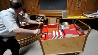 Ein Mann schiebt einen karren voll mit roten Büchern durch den Nationalratssaal.
