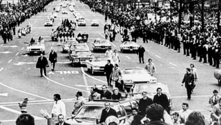 Wagenkolonne von Nixon auf einer grossen, menschengesäumten Strasse, aus dem vordersten Auot winken stehend Nixon und seine Gattin 