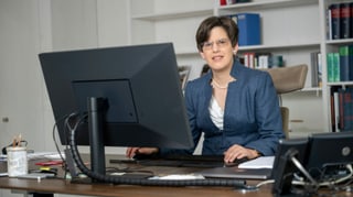 Frau Stüber sitzt in ihrem blauen Anzug am Schreibtisch. Vor ihr steht ein grosser Bildschirm und ein Telefon. Sie schaut in die Kamera. Frau Stüber sitzt in ihrem blauen Anzug am Schreibtisch. Vor ihr steht ein grosser Bildschirm und ein Telefon. Sie schaut in die Kamera.
