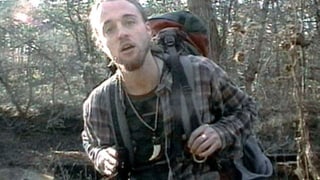 Ein junger Mann mit Rucksack steht im Wald und blickt in die Kamera.
