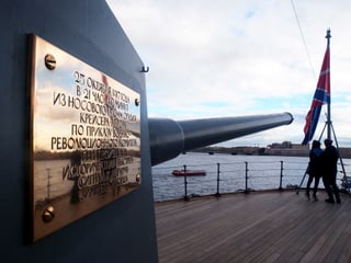 Das Kriegsschiff Aurora mit einer goldenen Aufschrift. Auf dem heutigen Museumsschiff stehen zwei Touristen. Das Kriegsschiff Aurora mit einer goldenen Aufschrift. Auf dem heutigen Museumsschiff stehen zwei Touristen.