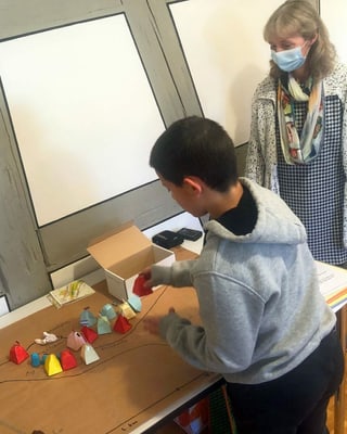 Ein Junge steht an einem Punlt und spielt mit farbigen Pyramiden, daneben steht eine Lehrerin mit Maske. Ein Junge steht an einem Punlt und spielt mit farbigen Pyramiden, daneben steht eine Lehrerin mit Maske.