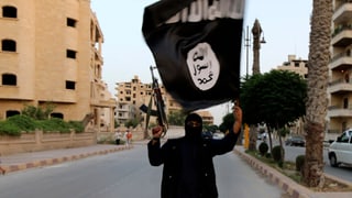 Ein schwarz gekleideter IS-Kämpfer mit Gesichtsmaske und erhobenem Maschinengewehr schwenkt eine IS-Flagge auf einer leeren Strasse.