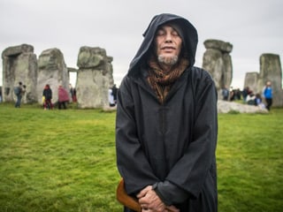Mann in schwarzem Umhang mit Kapuze steht mit geschlossenen Augen vor Steinkreis