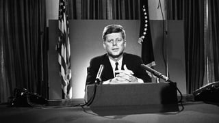 John F. Kennedy in einer TV-Ansprache vor US-Flagge.