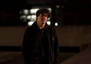 Mann in schwarzer Jacke steht nachts draussen, im Hintergrund unscharf Lichter eines Gebäudes