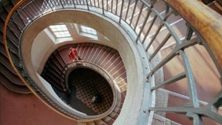 Eine Frau in rotem Pulli geht eine schneckenförmig gewundene Treppe hoch.