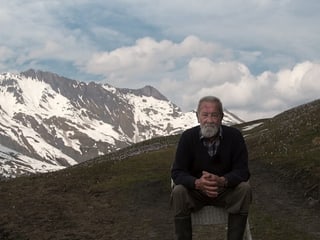 Mann sitzt auf einem Stuhl, im Hintergrund verschneite Berge.