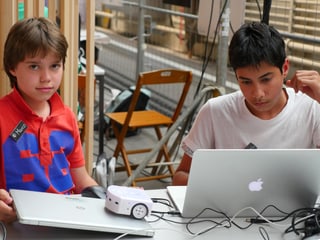 Zwei Jungen sitzen vor Laptops, auf dem einen, zugeklappten Laptop steht ein kleiner, weisser Roboter.