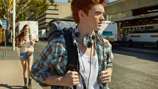 Ein junger Mann mit Rucksack und Kopfhörer läuft eine Strasse entlang, hinter ihm geht eine junge Frau ebenfalls mit Rucksack. Ein junger Mann mit Rucksack und Kopfhörer läuft eine Strasse entlang, hinter ihm geht eine junge Frau ebenfalls mit Rucksack.