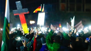 Eine Menschenmenge in der Nacht. Die Leute halten ihre Handys hoch. Im Bildmittelgrund hält ein Pegida-Demonstrant in Dresden ein mannshohes Kreuz in den Landesfarben in die Höhe.