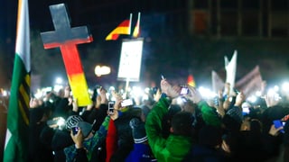Eine Menschenmenge in der Nacht. Die Leute halten ihre Handys hoch. Im Bildmittelgrund hält ein Pegida-Demonstrant in Dresden ein mannshohes Kreuz in den Landesfarben in die Höhe. Eine Menschenmenge in der Nacht. Die Leute halten ihre Handys hoch. Im Bildmittelgrund hält ein Pegida-Demonstrant in Dresden ein mannshohes Kreuz in den Landesfarben in die Höhe.