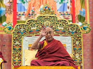 Dalai Lama auf einem grossen Sessel, er winkt.