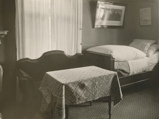 schwarz/weiss Bild von Zimmer mit Teppichboden kleinem Tisch und kleinem Bett