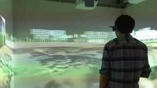 Mann steht in einem Raum. An den Wänden ist eine virtuelle Stadt projeziert. Er trägt eine 3D-Brille. 