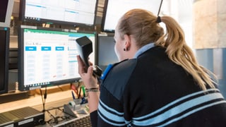 Eine Frau sitzt vor vielen Monitoren. Sie hält einen Telefonhörer in der Hand. Eine Frau sitzt vor vielen Monitoren. Sie hält einen Telefonhörer in der Hand.