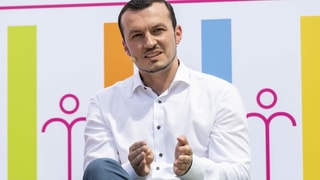 Ein Mann mit Mikrofon sitzt vor einer farbigen Plakatwand.