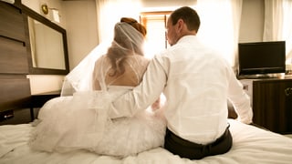 Hochzeitspaar in Hochzeitskleidung auf dem Bett, sitzend, Ansicht von hinten