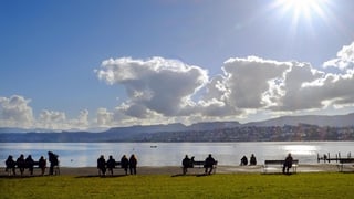 Menschen sitzen auf Bänken am Zürichsee bei strahlendem Sonnenschein.