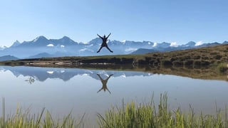 Eine Frau springt in die Luft vor Alpenpanorama. Davor ein See. 