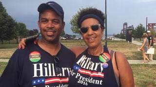 Eine Afroamerikanerin mit ihrem Arm um einen Afroamerikaner. Beide tragen Hillary Clinton for President T-Shirts.
