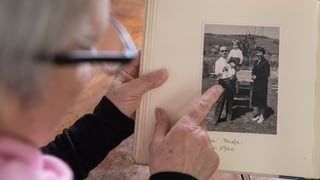 Eine Frau zeigt auf ein offenes Fotoalbum. Zu sehen ist eine Schwarz-Weiss-Aufnahme eines kleinen Kindes auf dem Arm eines Mannes. Daneben steht eine Frau. 