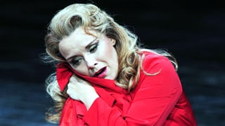 Eine Frau mit blonden Locken sitzt auf der Bühne, die Arme um sich geschlungen, der Blick leidend. 