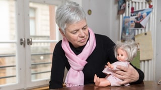 Eine Frau sitzt an einem Tisch. Sie schaut auf eine Puppe, die sie in der Hand hält. 