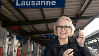 Pilloud lächelt mit einem Handy in der Hand in die Kamera, oberhalb von ihr das Bahnhof-Schild von Lausanne.