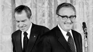 Schwarzweissbild: Kissinger steht grinsend vor einem Mikrofon. Hinter ihm geht Richard Nixon.