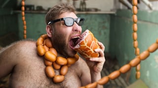 Ein behaarter Mann mit Sonnenbrille beist in ein Stück Fleisch.