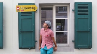 Mann sitzt auf Fensterbank, über ihm ein Wegweiser «Huberweg» Mann sitzt auf Fensterbank, über ihm ein Wegweiser «Huberweg»