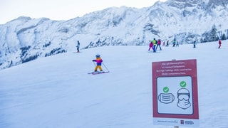 Kind fährt Ski vor Schild, auf dem die Maskenpflicht visualisiert ist.