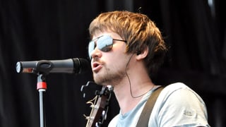 Vasya Oblomov, mit Sonnenbrille und Gitarre, singend am Mikrofon.
