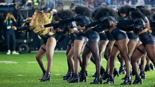 Beyoncé singt auf einem Rasen. Hinter ihr zahlreiche Frauen in schwarzem, knappen Lederkostüm.