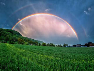 Dunkle Wolken, vor denen ein Regenbogen mit Doppelbogen leuchtet. Unten satt grünes Feld. 