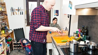 Ein Mann kocht in der Küche, die Frau sitz mit Baby am Tisch