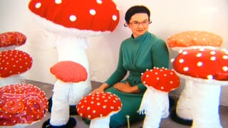 Ivana Falconi in einem grünen Kleid zwischen vielen, unterschiedlich grossen Fliegenpilzen aus Stoff und andferen Materialien.