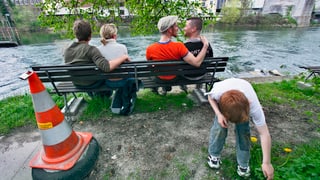 Ein hetero- und ein homosexuelles Paar sitzen auf einer Bank, davor spielt ein Kind. 