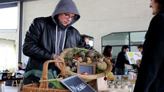 Ein Mann mit schwarzer Jacke und Kapuze hält einen Gemüsekorb in den Händen.