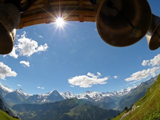 Oben im Bild ein Balken mit Glocken, die Sonne scheint unter dem Balken durch. In der Mitte ist blauer Himmel und unten das Alpenpanorama mit Schneebergen. 