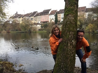 Adrian Küpfer und Sabine Dahinden schauen hinter einehm Baum hervor. Im Hintergrund sieht man eine Stadt. 